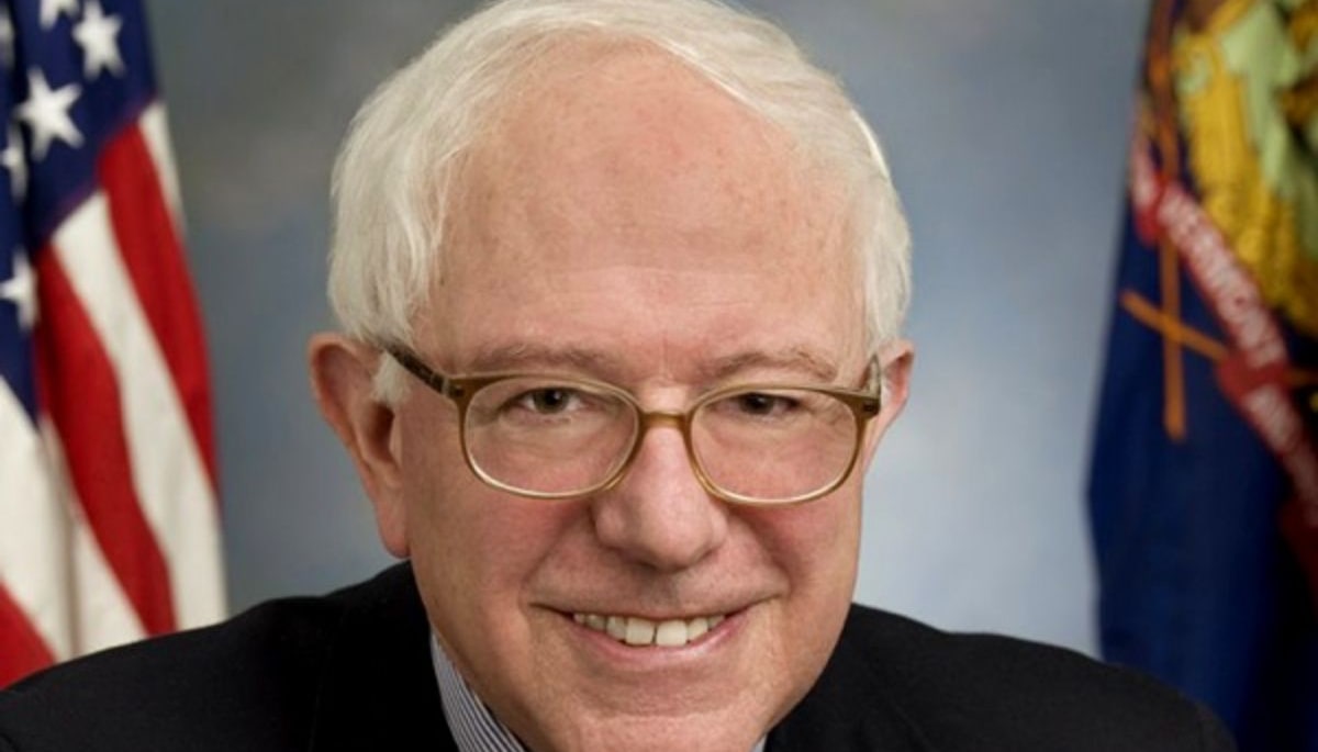Bernie Sanders smiling in his official U.S. Senate portrait, wearing glasses and a dark suit with a red patterned tie, flanked by the American and Vermont flags.