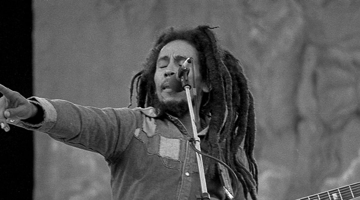 Bob Marley singing into a microphone stand at Dalymount Park, Dublin, on his final tour in July 1980, dreadlocks loose around his face and one arm outstretched toward the crowd.