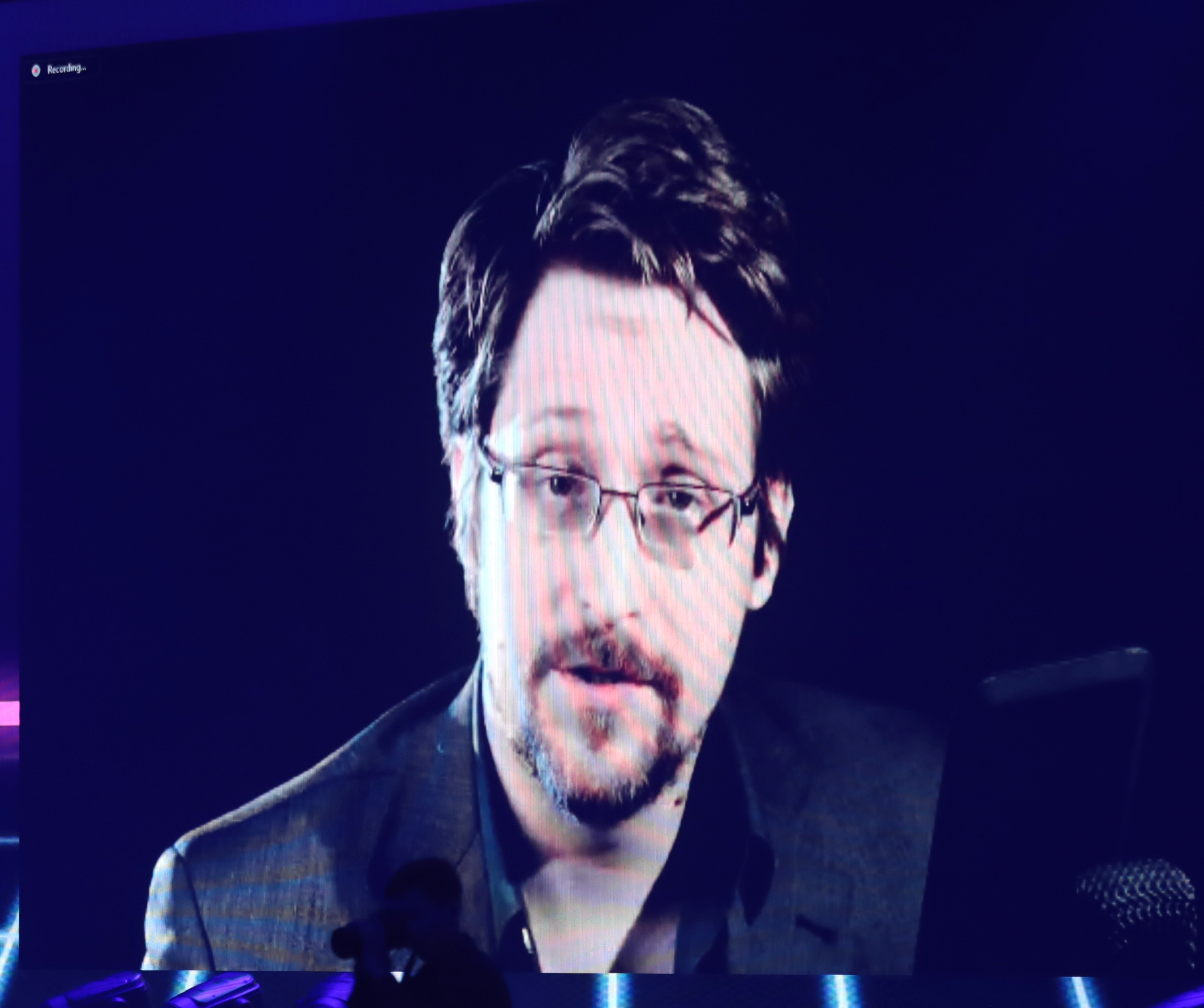 Edward Snowden appearing by video link on a large screen at a 2021 conference, head and shoulders, wearing glasses and a gray blazer.