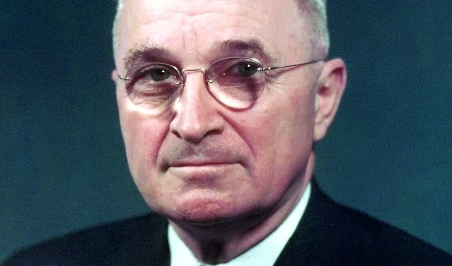 Harry S. Truman in a color portrait, wearing round wire-rimmed glasses and a dark suit.