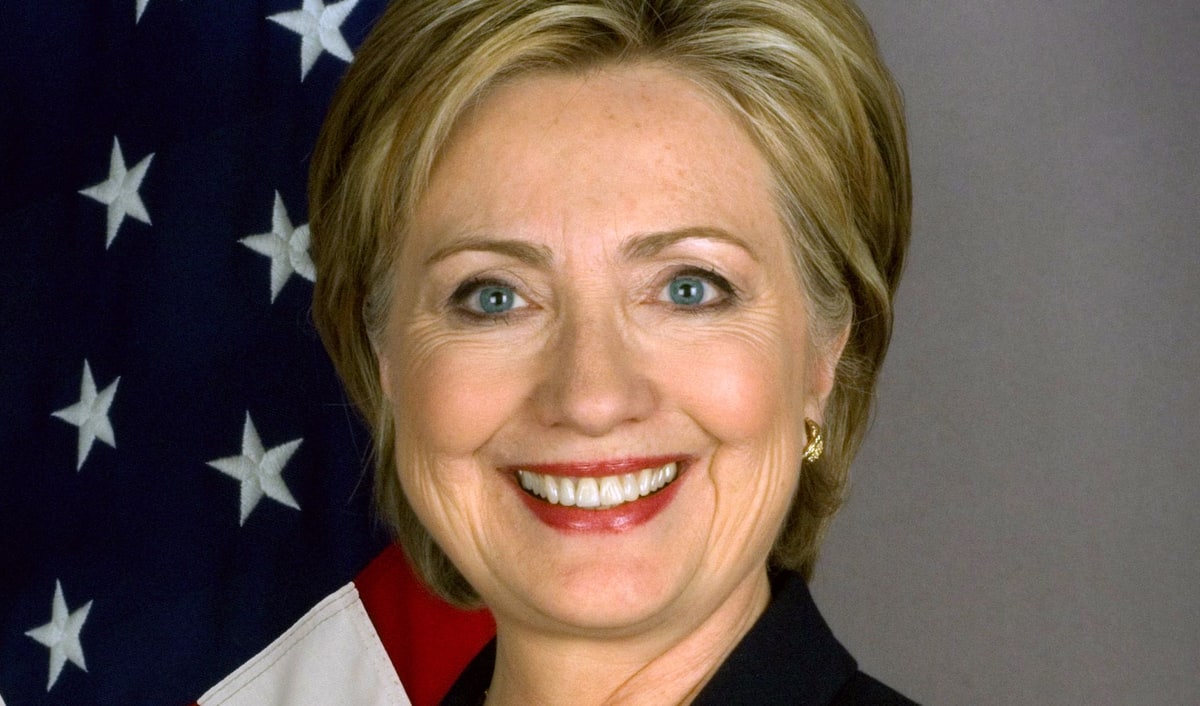 Hillary Clinton smiling in her official U.S. Department of State portrait as Secretary of State, standing before an American flag in a dark blazer and orange top.