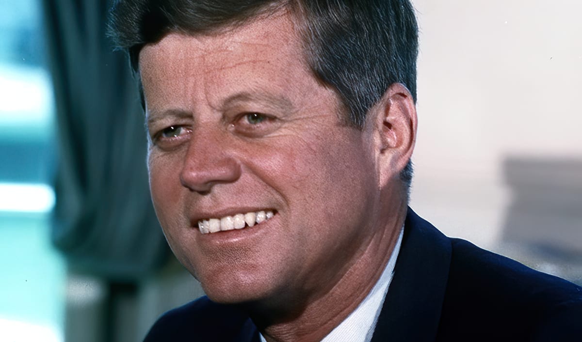 John F. Kennedy in his White House color portrait, smiling in a dark suit.