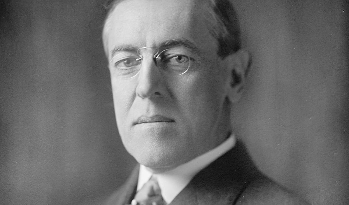 Woodrow Wilson in a photographic portrait, wearing pince-nez glasses and a dark suit.